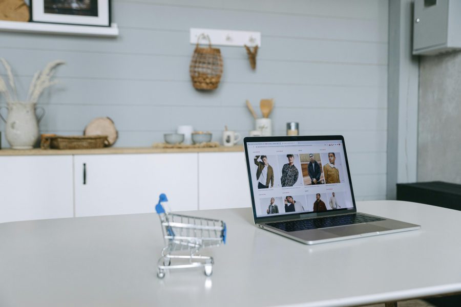 A laptop displaying an online shopping portal sits on a minimal desk with a miniature shopping cart in focus.